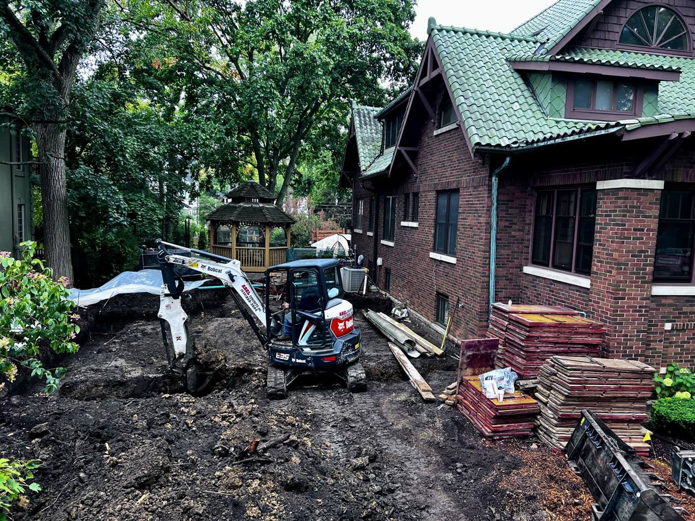 Mini excavator digging behind a brick bungalow with a green tile roof, a gazebo in the background