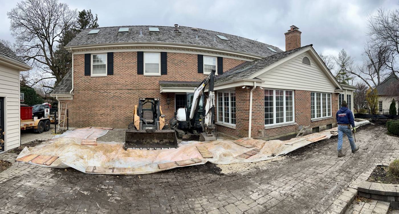 Back of a brick colonial in Wheaton with plastic and boards over the paver patio and a skid steer and mini excavator arriving
